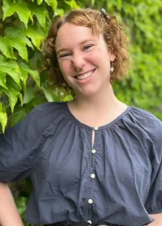 Mental health counselor Alex Chapman with curly red hair and a septum piercing, smiling in front of a wall covered in green ivy.