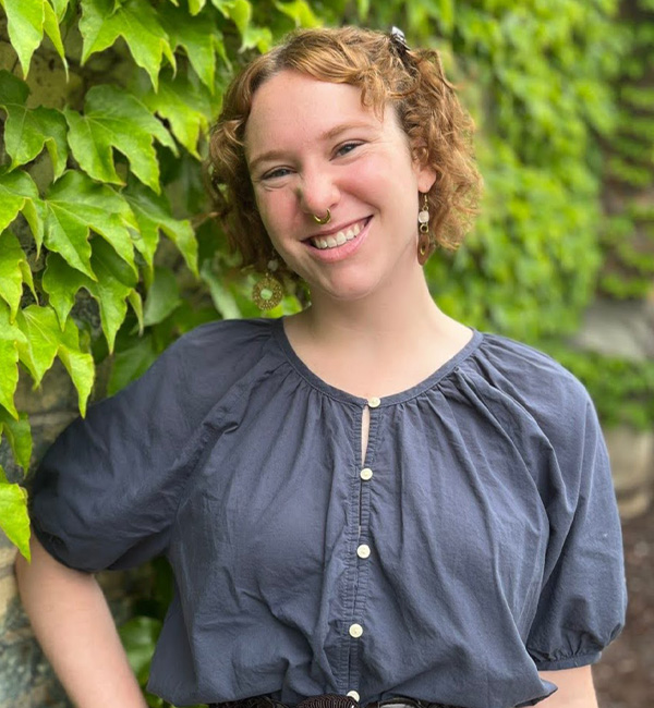 Mental health counselor Alex Chapman with curly red hair and a septum piercing, smiling in front of a wall covered in green ivy.