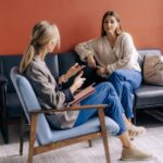 Two women sit in a room having a serious conversation; one gestures while holding a notebook, and the other listens attentively on a couch with a bag beside her, reflecting the focused atmosphere of a therapy or psychotherapy session addressing anxiety.