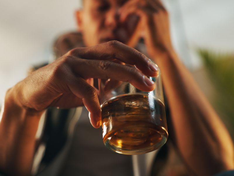 A person holding a glass of whiskey with ice, their face blurred in the background, and their other hand touching their face in a contemplative gesture, suggests an intimate moment often associated with anxiety or moments of self-reflection that can arise before or after therapy.
