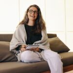 A smiling woman with long hair and glasses sits on a sofa, holding a notebook and pen—perhaps preparing for a therapy or psychotherapy session. She wears a light shawl, black shirt, and white pants, appearing relaxed and happy in a bright, modern setting.