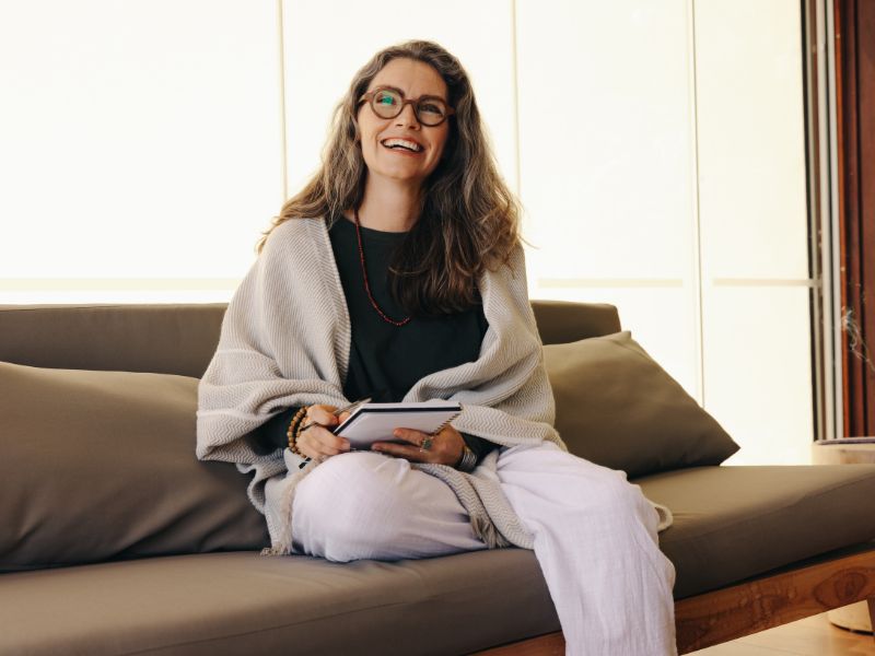 A smiling woman with long hair and glasses sits on a sofa, holding a notebook and pen—perhaps preparing for a therapy or psychotherapy session. She wears a light shawl, black shirt, and white pants, appearing relaxed and happy in a bright, modern setting.