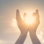 Two hands raised toward a bright sun, with sunlight streaming between the fingers, against a soft, blurred sky—a gentle image that evokes hope and the healing journey often found in psychotherapy for anxiety or depression.
