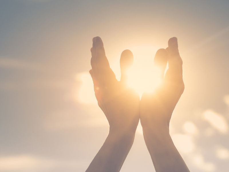 Two hands raised toward a bright sun, with sunlight streaming between the fingers, against a soft, blurred sky—a gentle image that evokes hope and the healing journey often found in psychotherapy for anxiety or depression.