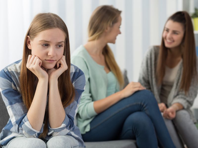 A girl sits alone off to the side of a group of others, demonstrating social anxiety.