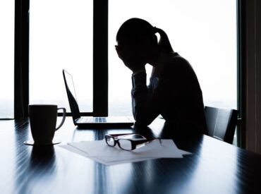 A woman is silhouetted in front of large office windows with her face in her hands while sitting in front of her laptop.