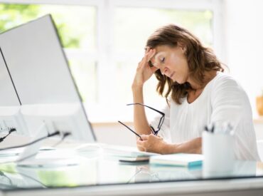 A woman in a white shirt sits at a desk holding her glasses, appearing stressed or tired, with her hand on her forehead in front of a computer monitor—signs that may suggest she is struggling with depression and could benefit from therapy.