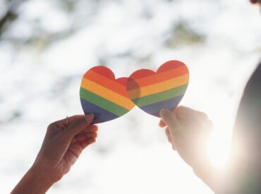 An image of two hands holding LGBTQ rainbow hearts together