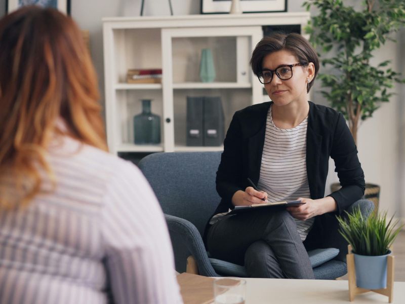 A person with short hair and glasses sits in a chair, holding a clipboard and pen, attentively listening to another person in an office setting with plants and shelves in the background—creating a supportive environment for therapy sessions focused on depression or anxiety.