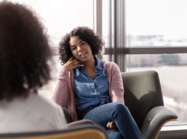 A woman with curly hair sits comfortably in a chair, smiling and talking to another person in a bright room with large windows, engaging in therapy to address anxiety and depression.