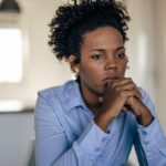 A woman wearing a light blue shirt sits indoors with her hands clasped under her chin, looking thoughtful and concerned, as if reflecting on her experiences with depression or anxiety.