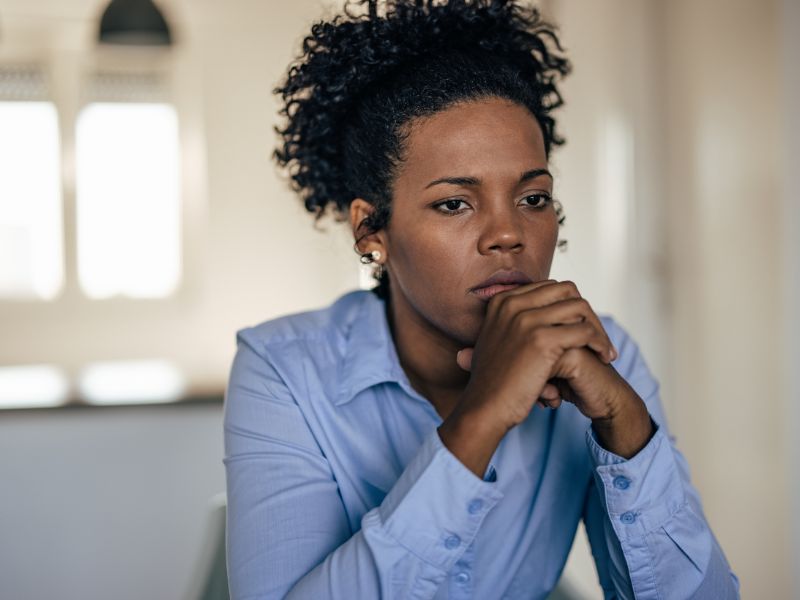 A woman wearing a light blue shirt sits indoors with her hands clasped under her chin, looking thoughtful and concerned, as if reflecting on her experiences with depression or anxiety.