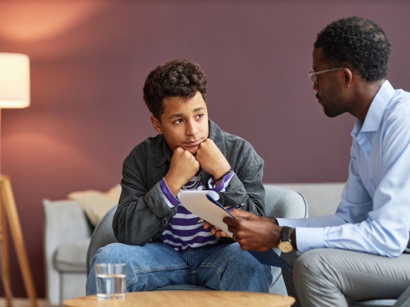 A teenage boy sits with his chin on his hands, looking thoughtful, while an adult man holding a clipboard talks to him in a cozy room during a therapy session for depression and anxiety.