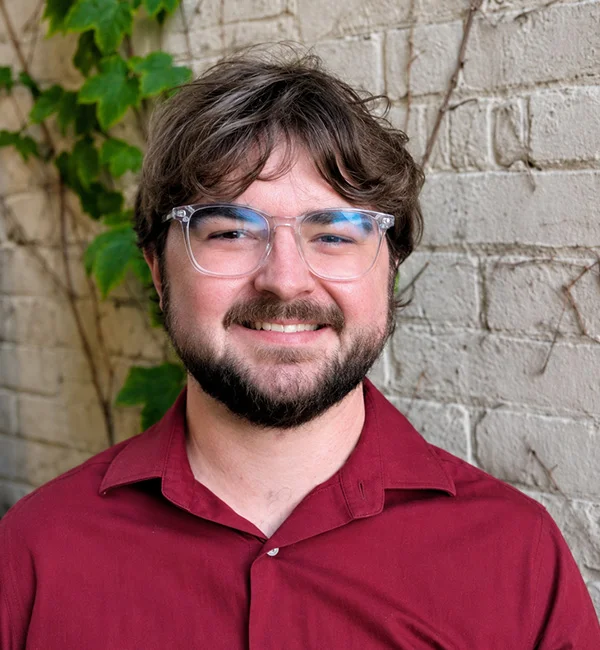 Nick Schiller, a person with brown hair, a beard, and glasses, smiles while standing in front of a white brick wall with green leaves growing on it. He is wearing a maroon shirt.